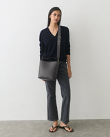 Person standing with shoulder bag, wearing a sweater and jeans, in sandals.