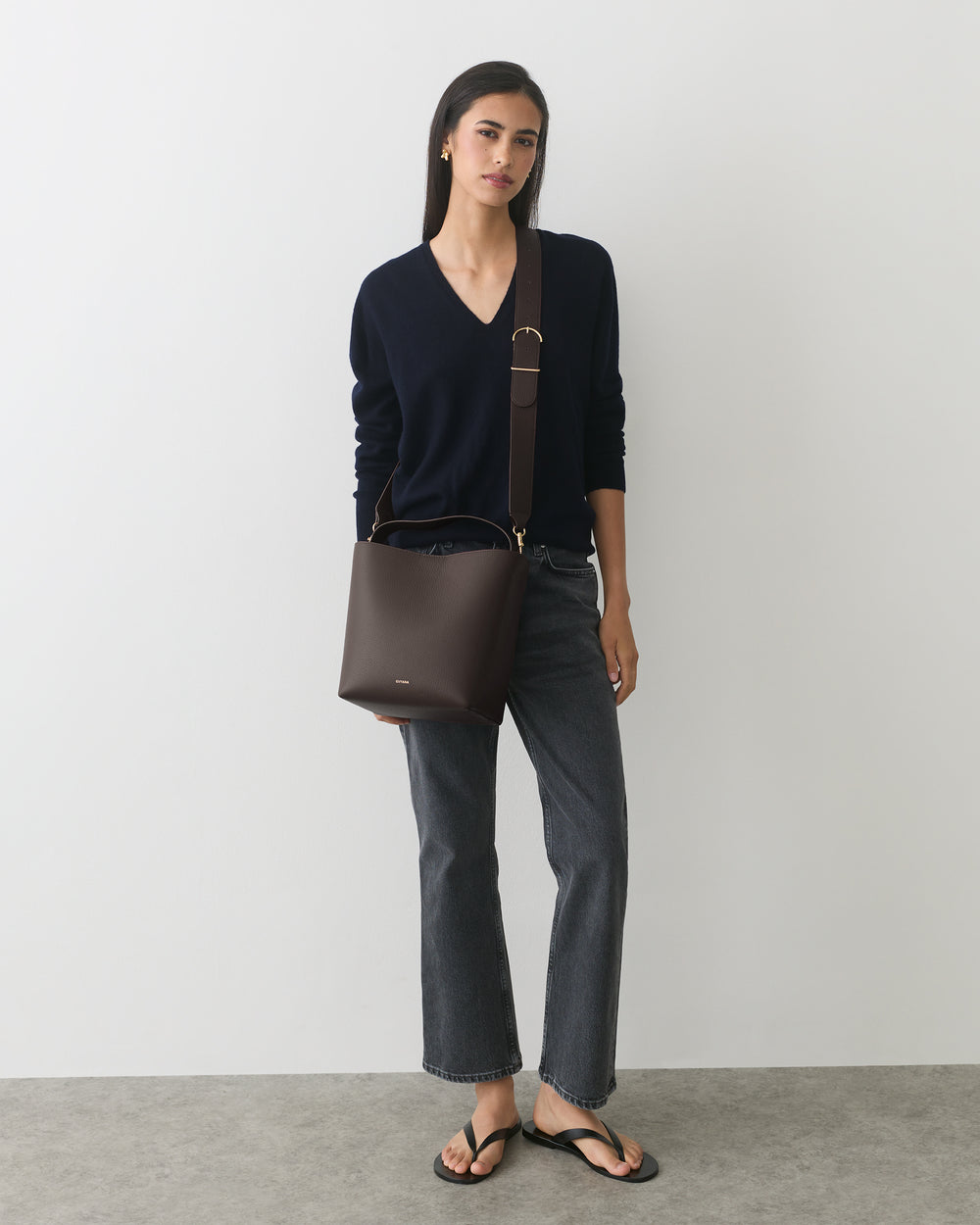 Person wearing sweater and jeans, holding shoulder bag, standing against plain background.