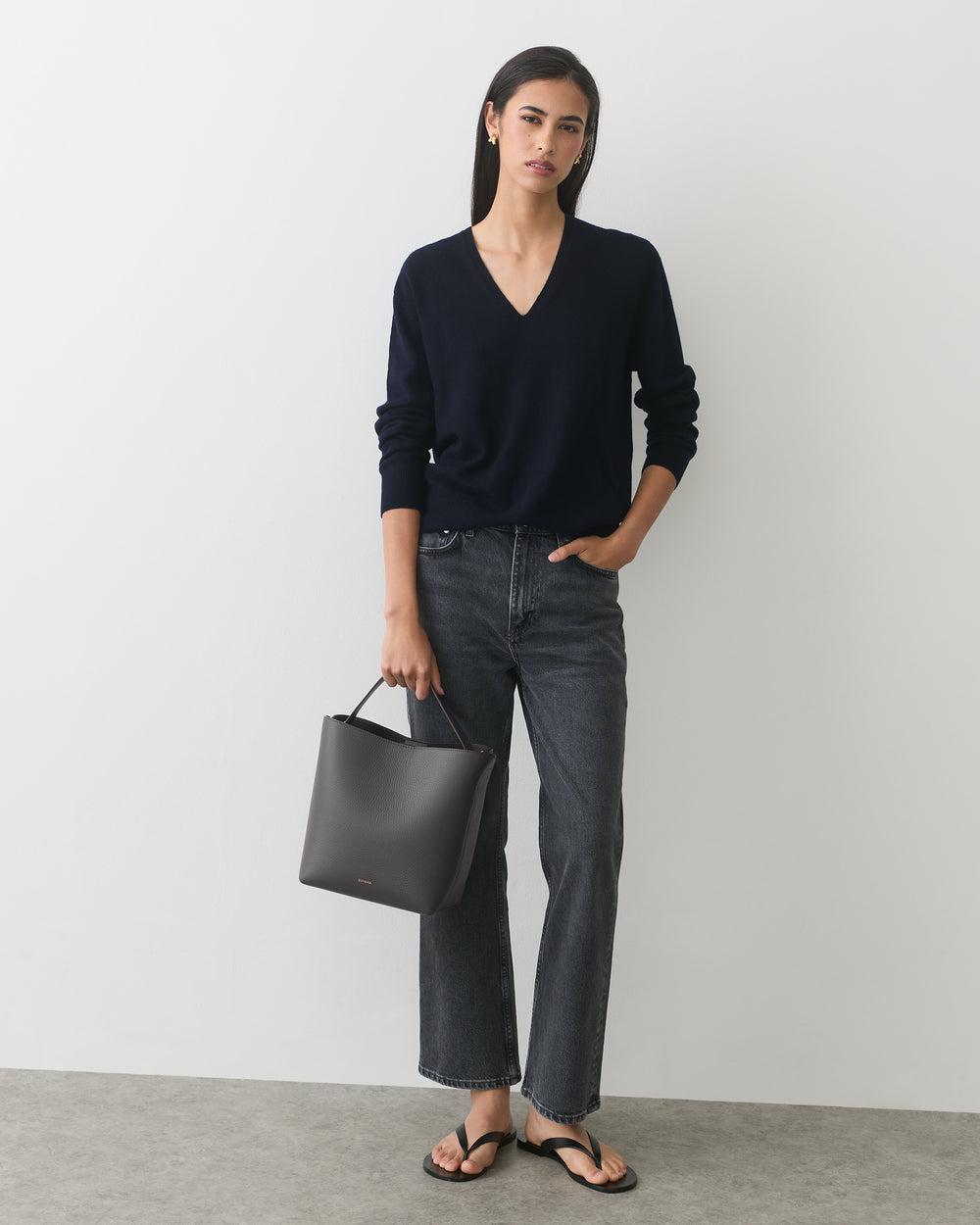 Person wearing sweater and jeans, holding a handbag, standing against plain background.