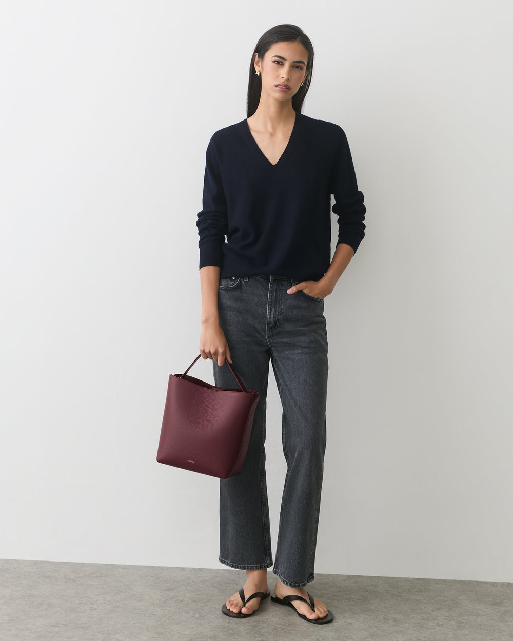 Person wearing a sweater and jeans holding a handbag, standing against a plain background.