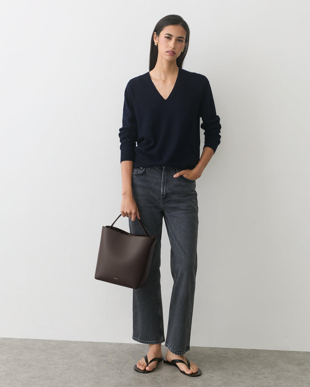 Person wearing a sweater and jeans holding a handbag, standing against a plain backdrop.