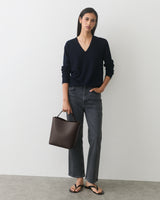 Person wearing a sweater and jeans holding a handbag, standing against a plain backdrop.