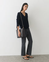 Person in a sweater and jeans holding a shoulder bag standing indoors.
