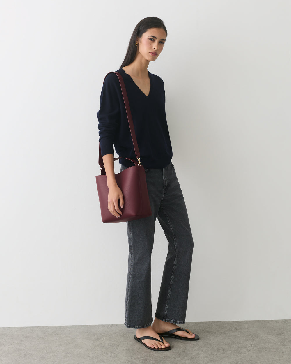 Person wearing a long-sleeve shirt and pants, holding a shoulder bag, stands against a plain wall.
