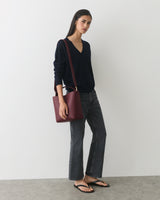 Person wearing a long-sleeve shirt and pants, holding a shoulder bag, stands against a plain wall.