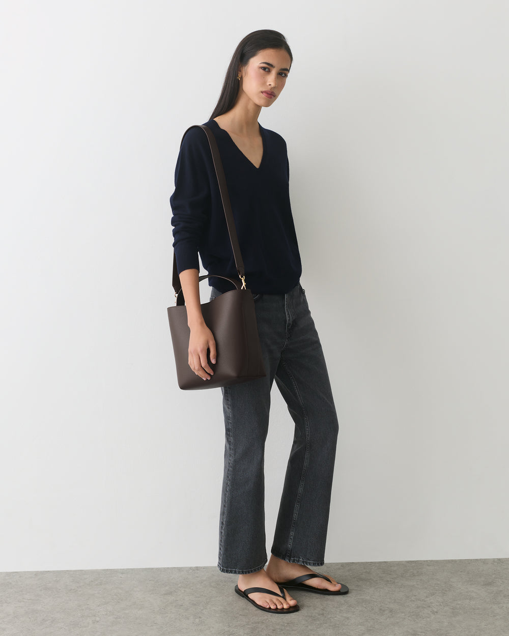 Person wearing sweater and jeans holding a shoulder bag, standing in a neutral setting.