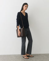 Person wearing sweater and jeans holding a shoulder bag, standing in a neutral setting.