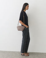 Person in casual outfit with small shoulder bag stands against white background.