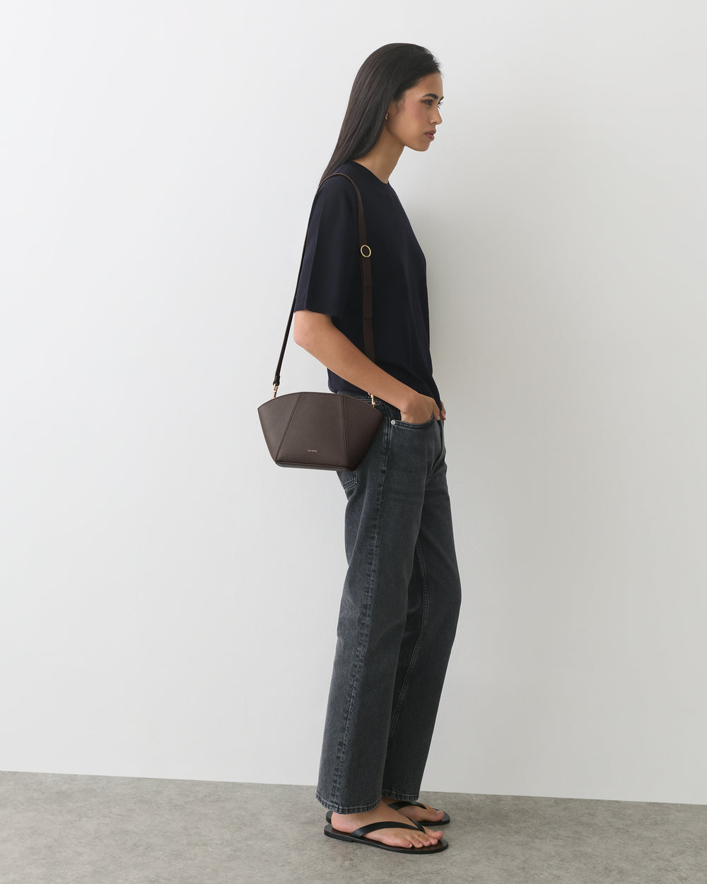 Person standing sideways, wearing a T-shirt, jeans, sandals, and carrying a small shoulder bag.