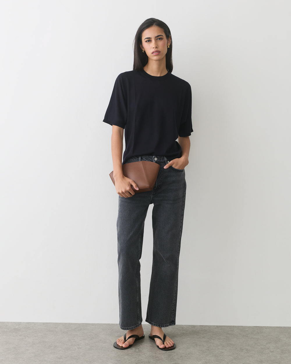 Person standing against wall holding a clutch wearing a t-shirt, jeans, and flip-flops