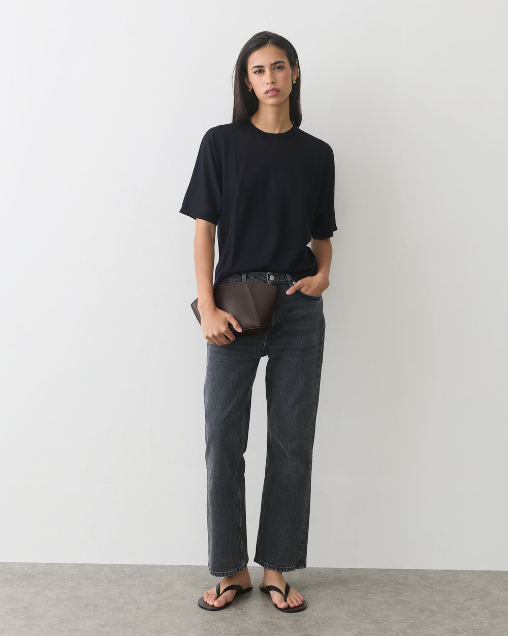 Person wearing jeans and t-shirt, holding a clutch, standing against plain background.