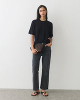 Person wearing jeans and t-shirt, holding a clutch, standing against plain background.