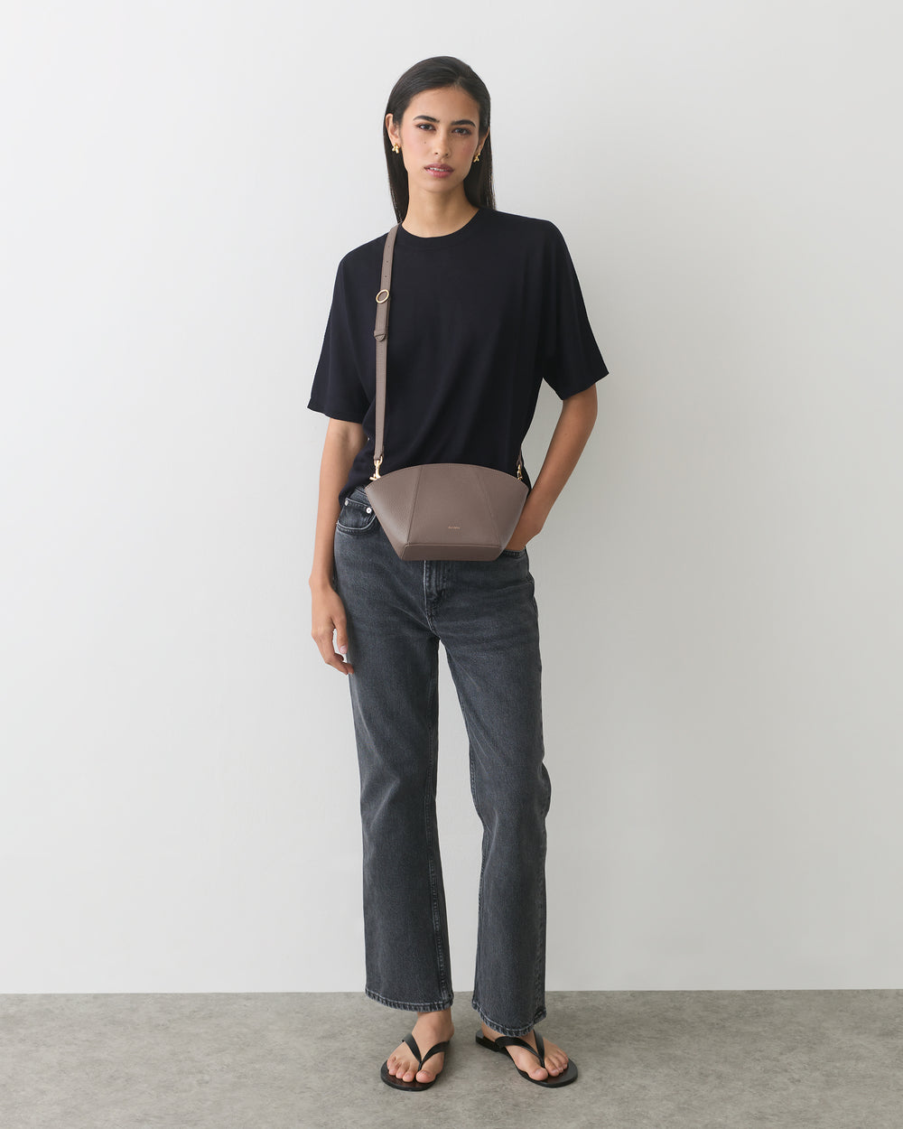 Person standing against a wall wearing a t-shirt, jeans, sandals, and a shoulder bag.