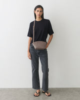 Person standing against a wall wearing a t-shirt, jeans, sandals, and a shoulder bag.