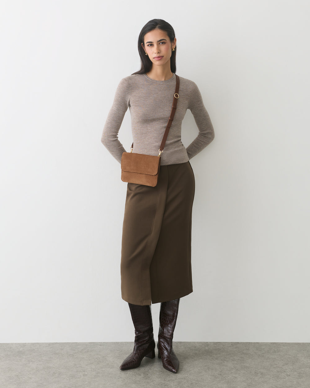 Person wearing sweater, long skirt, boots, and a crossbody bag standing against plain background.