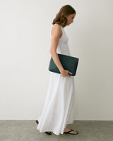 Person in long dress holding a clutch bag, wearing sandals, stands against plain backdrop.