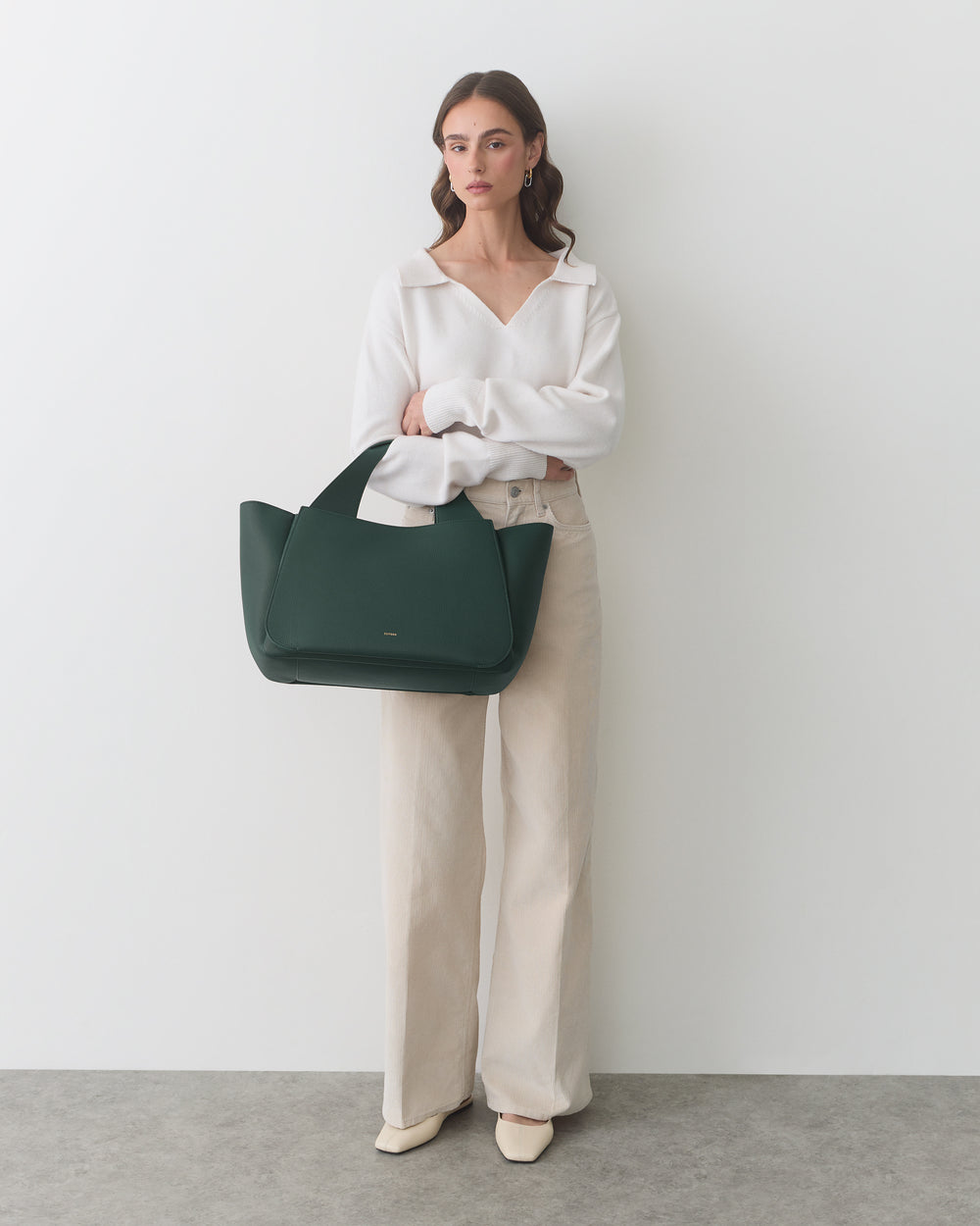Person standing with a travel bag, wearing long-sleeve top and wide-leg pants.