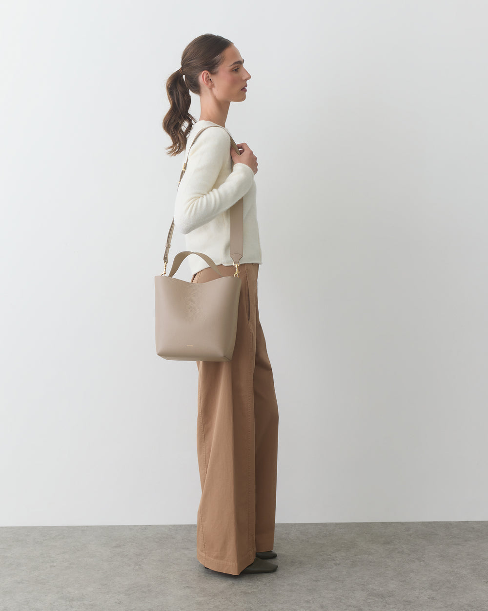 Person standing in profile holding a handbag, dressed in a sweater and wide-leg pants.