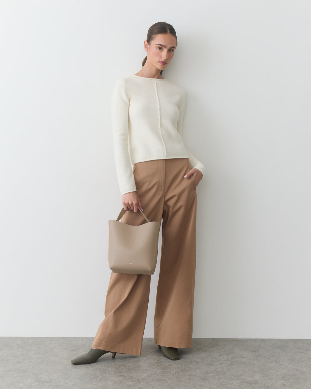 Person standing against wall holding a handbag, wearing a sweater and wide-leg pants.
