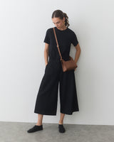 Person wearing a t-shirt and wide-leg pants with a shoulder bag leans against a wall
