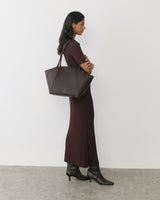Person in long dress carrying a large shoulder bag, side view against plain background.