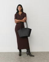 Person in a long dress holds a large tote bag, standing against a plain background.