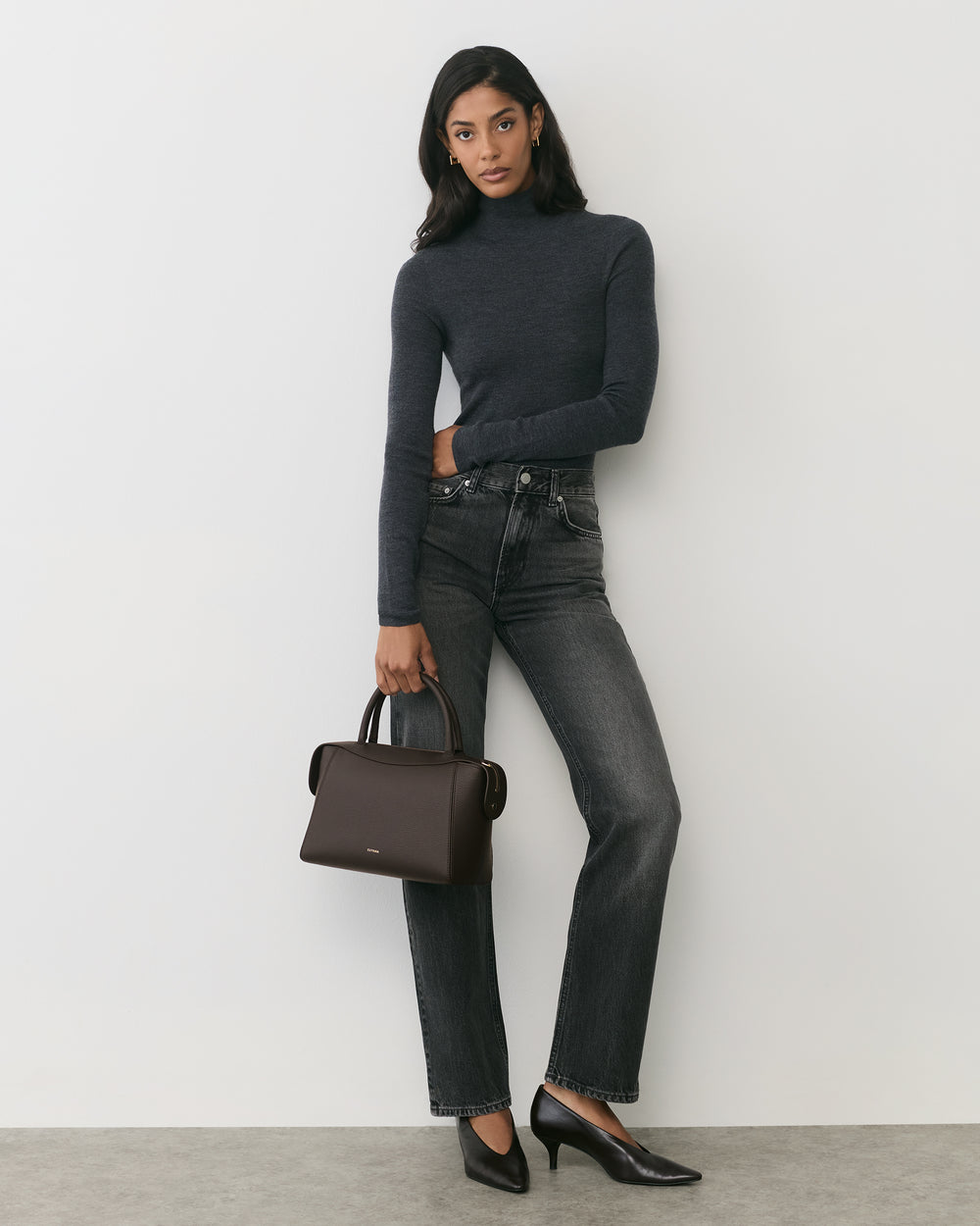 Person standing against wall holding a handbag, wearing a long-sleeve top and jeans.