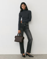 Person standing against wall holding a handbag, wearing a long-sleeve top and jeans.
