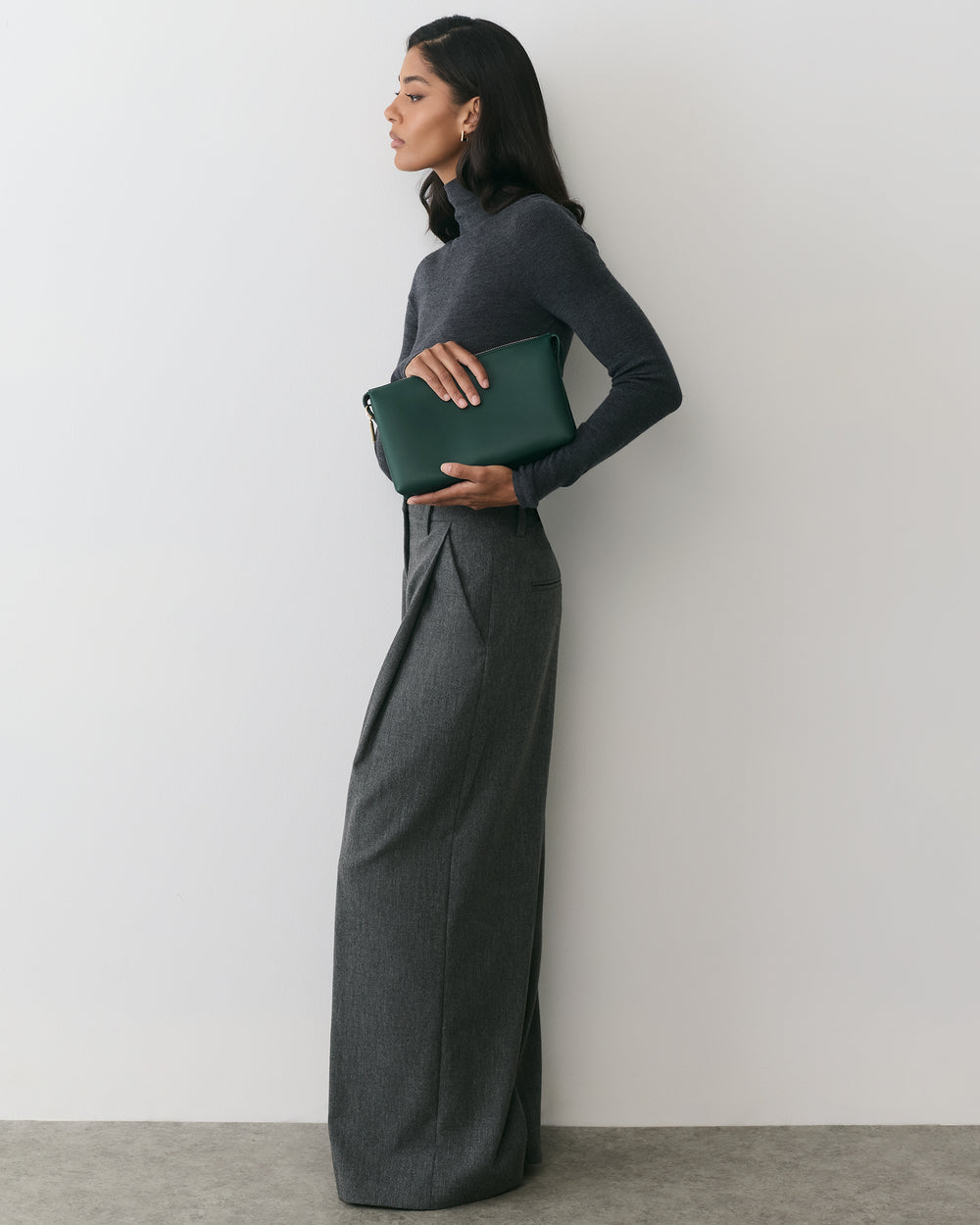 Person standing against a wall holding a clutch, wearing a long-sleeve top and wide-leg trousers.