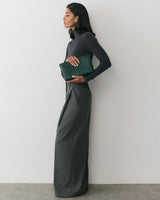 Person standing against a wall holding a clutch, wearing a long-sleeve top and wide-leg trousers.