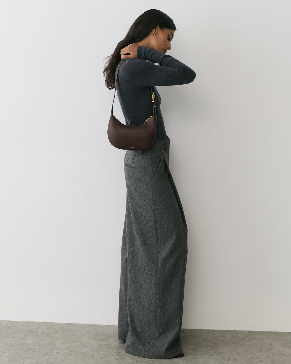 Person wearing long pants and sweater, holding a shoulder bag, standing sideways against a wall.