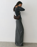 Person wearing long pants and sweater, holding a shoulder bag, standing sideways against a wall.