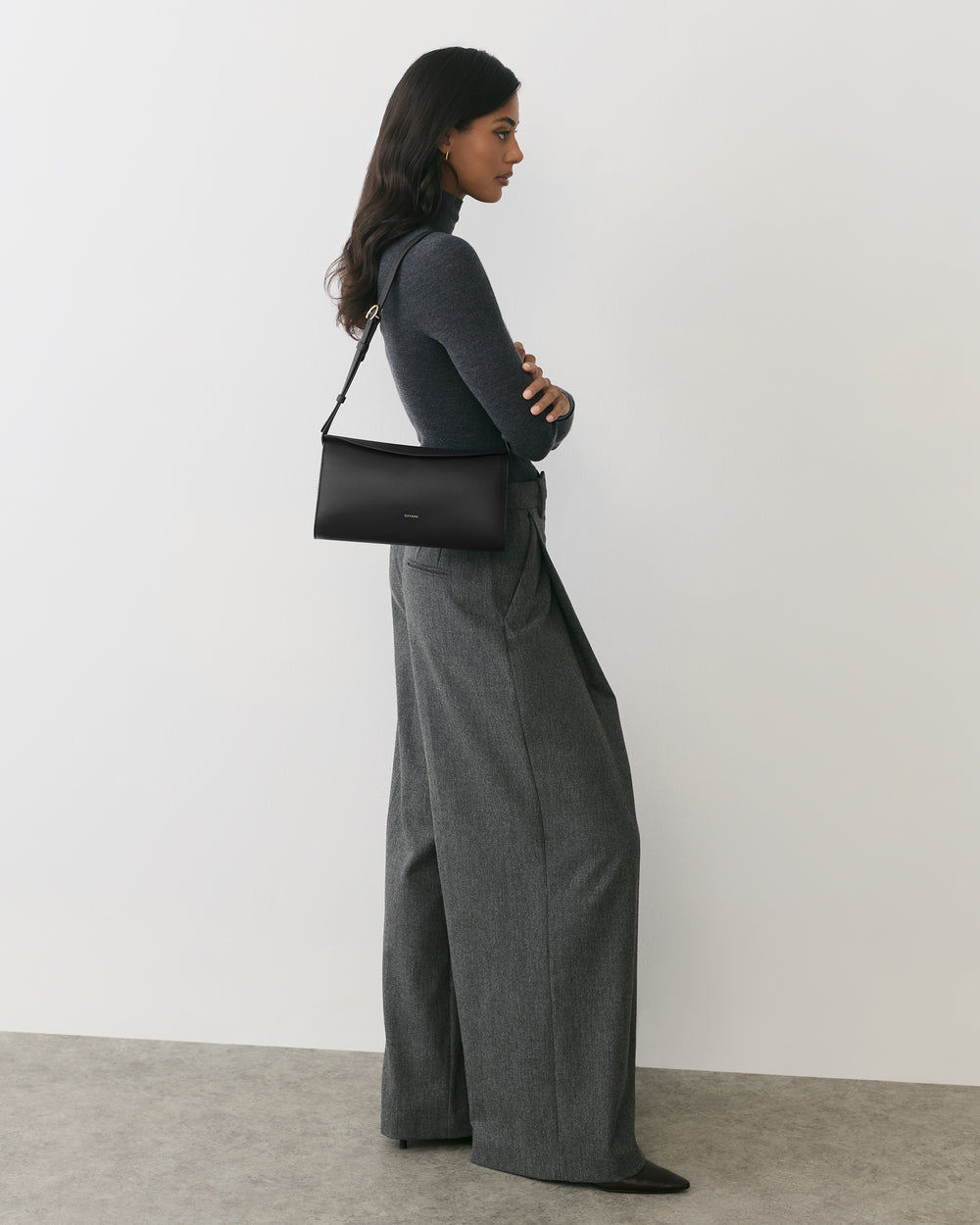 Person in long pants and top, holding a handbag, standing against a plain background.