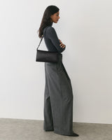 Person in long pants and top, holding a handbag, standing against a plain background.