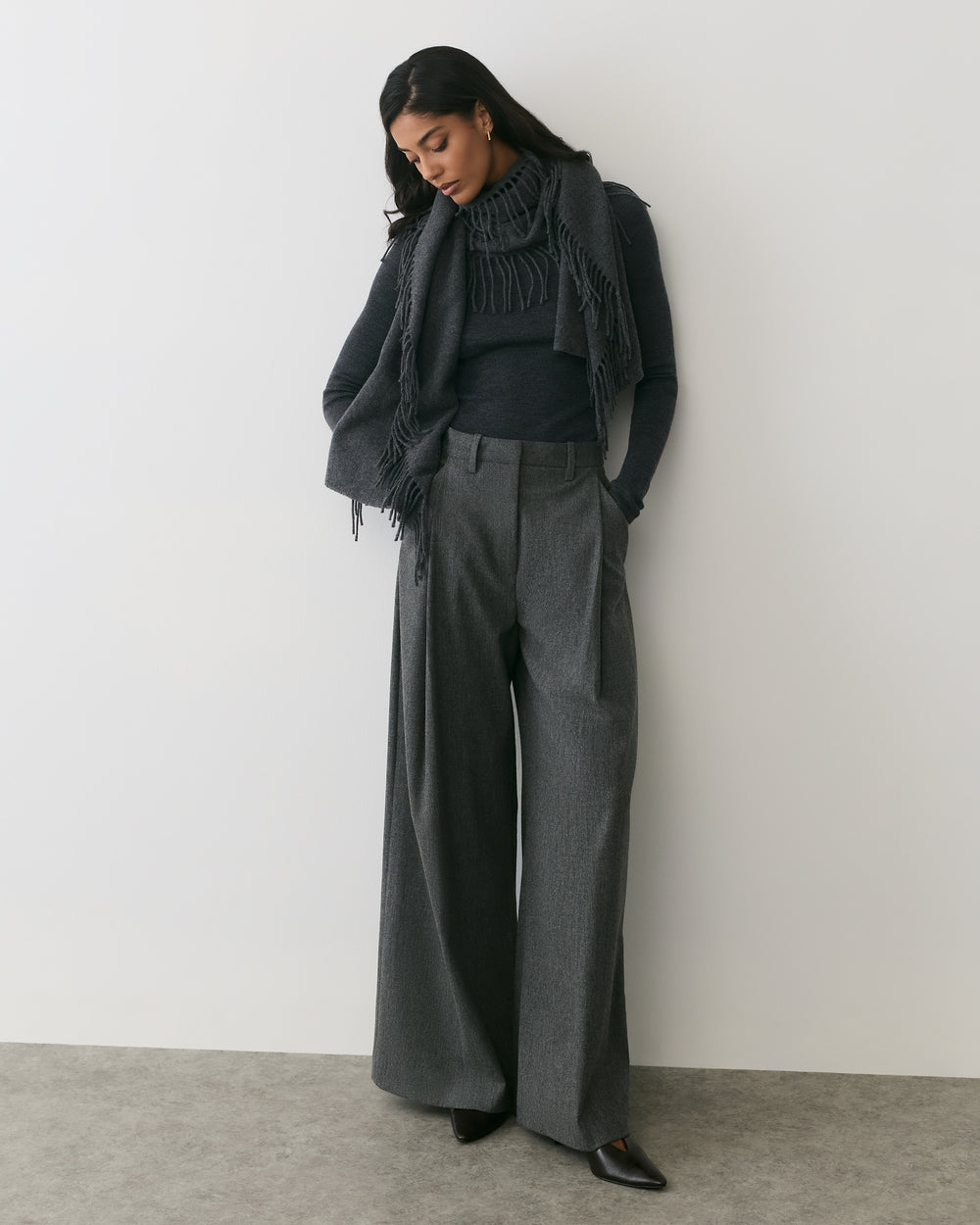 Person wearing wide-leg pants, sweater, and scarf stands against a plain wall.