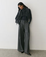 Person wearing wide-leg pants, sweater, and scarf stands against a plain wall.