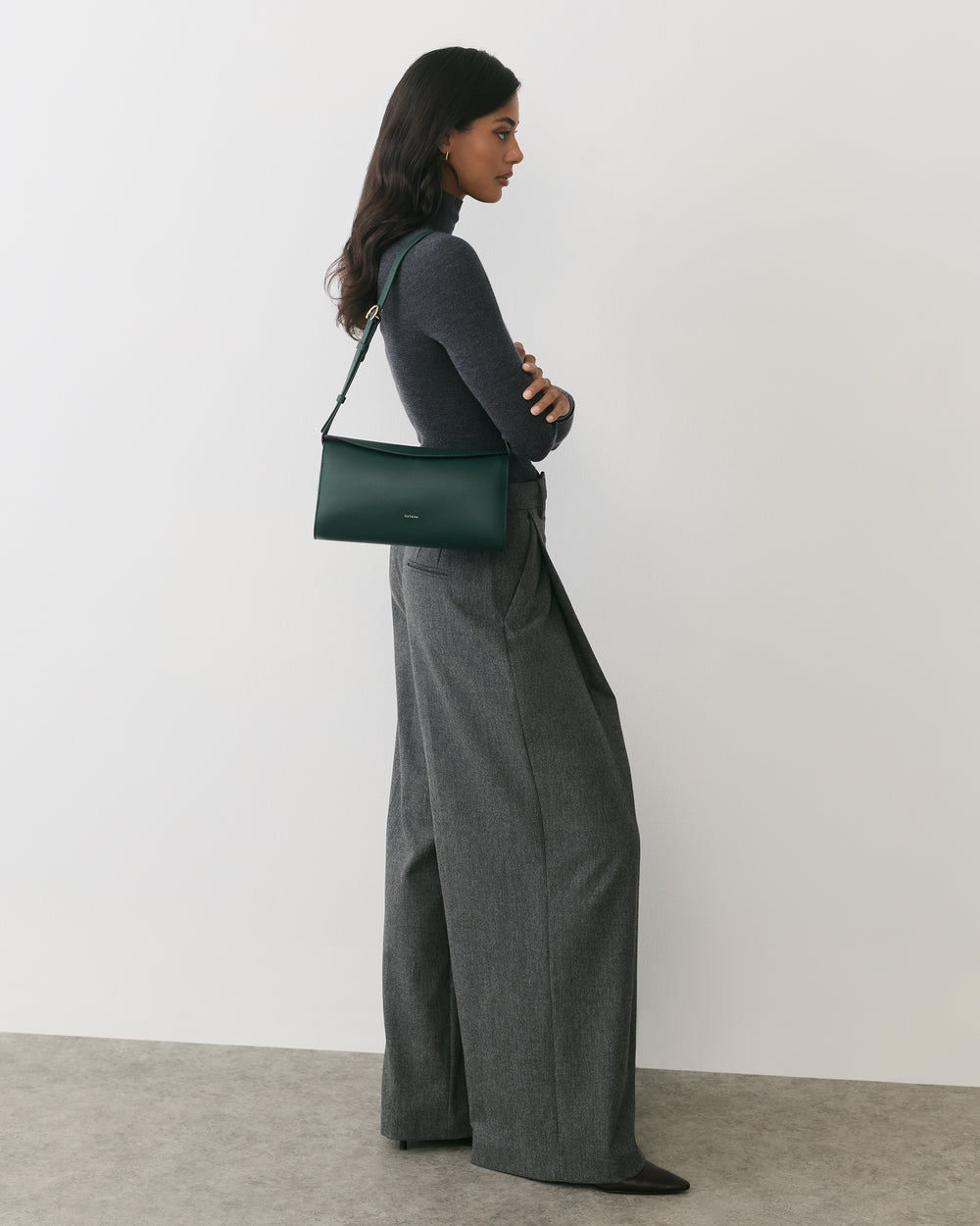 Person in long pants with shoulder bag standing against plain backdrop.