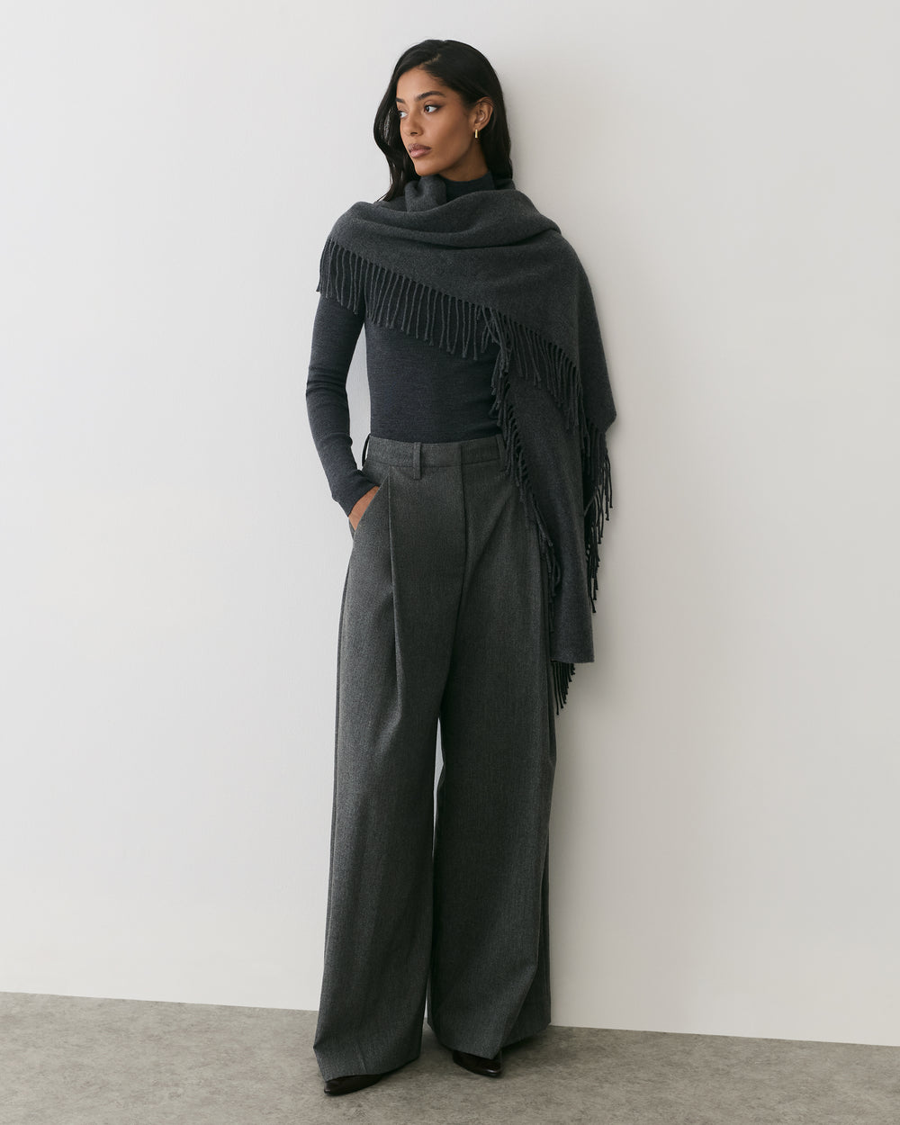 Person wearing a scarf wrap and wide-leg pants standing in front of a plain wall.
