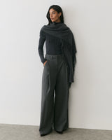 Person wearing a scarf wrap and wide-leg pants standing in front of a plain wall.