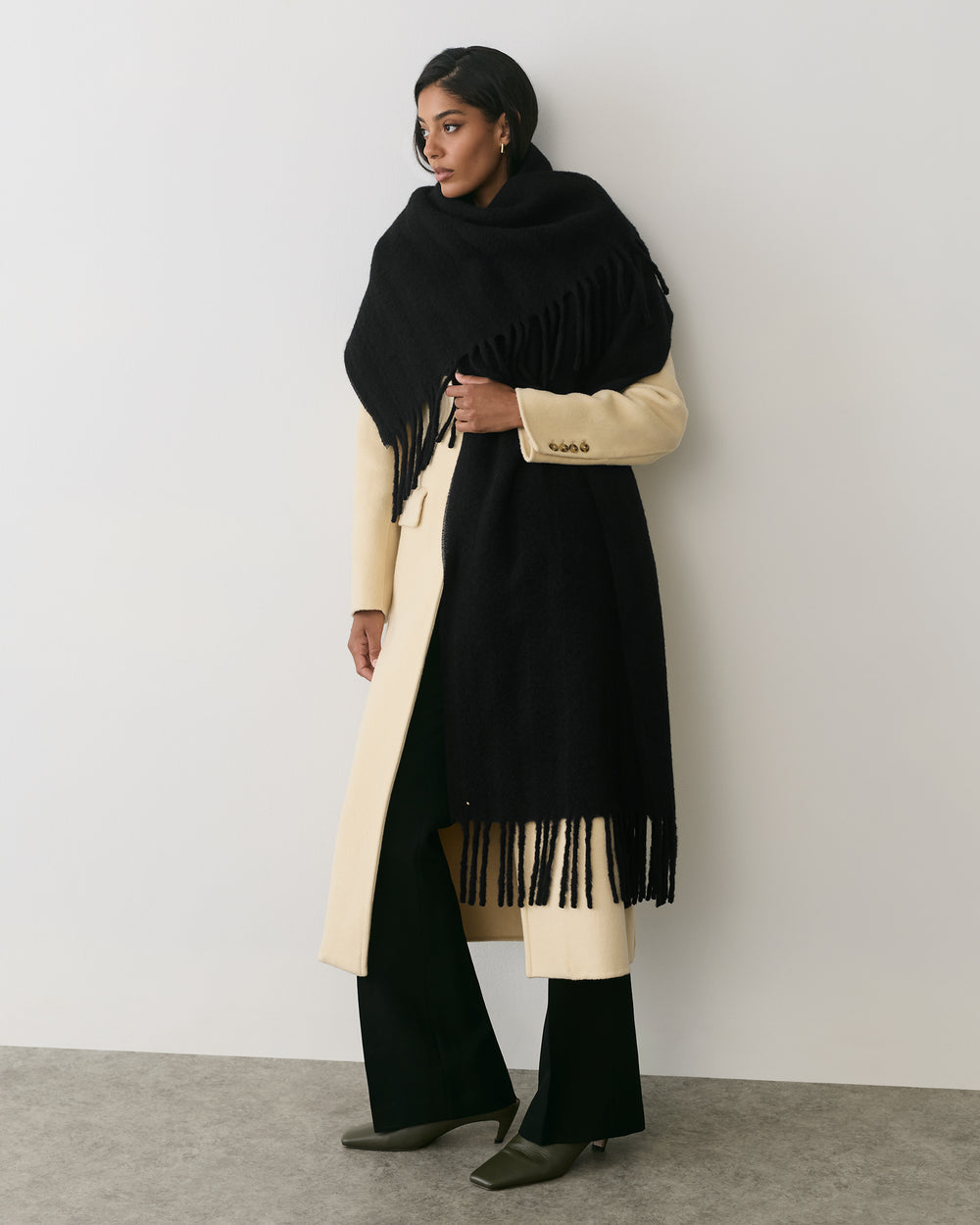 Person wearing long coat with large fringed scarf standing against plain background