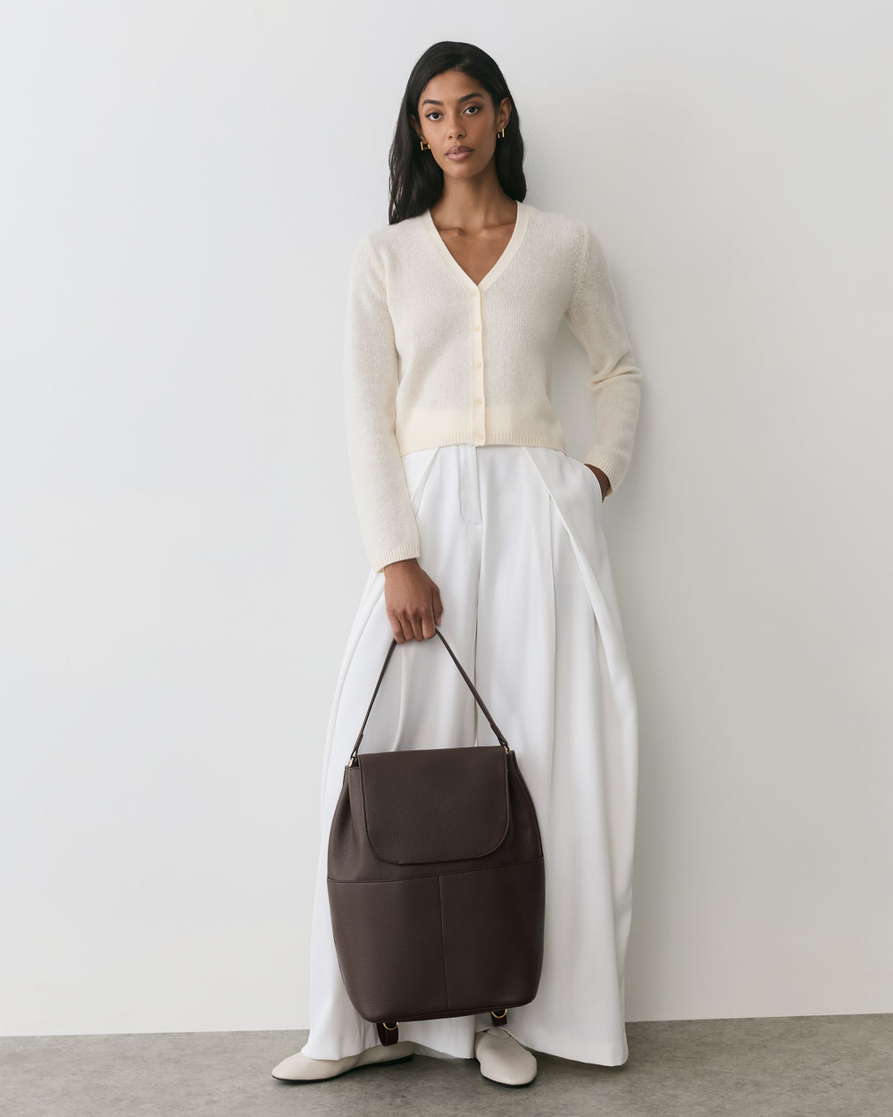 Person standing against wall holding large bag, wearing long skirt and cardigan