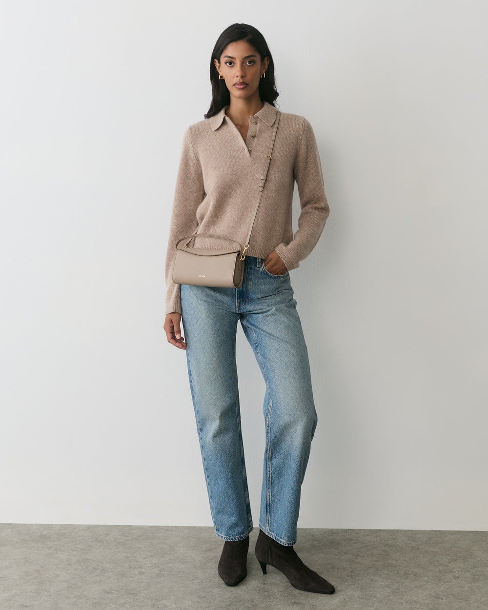 Person wearing a sweater, jeans, boots, and a shoulder bag, standing against a plain background.