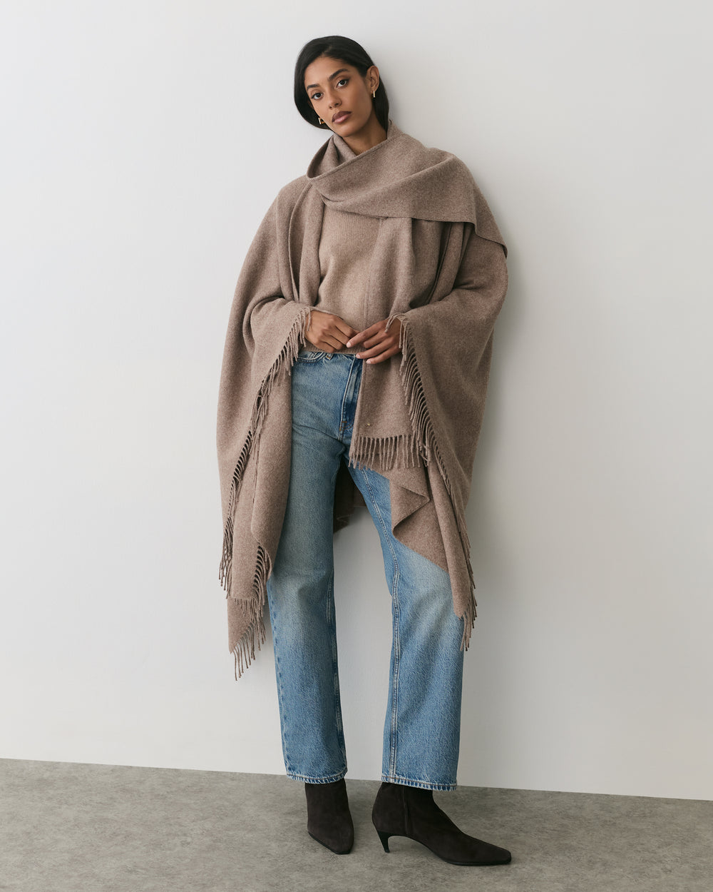 Person wearing a large fringed cape and jeans standing against a plain wall.