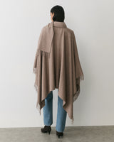 Person wearing cape over jeans with heels, standing against plain background.