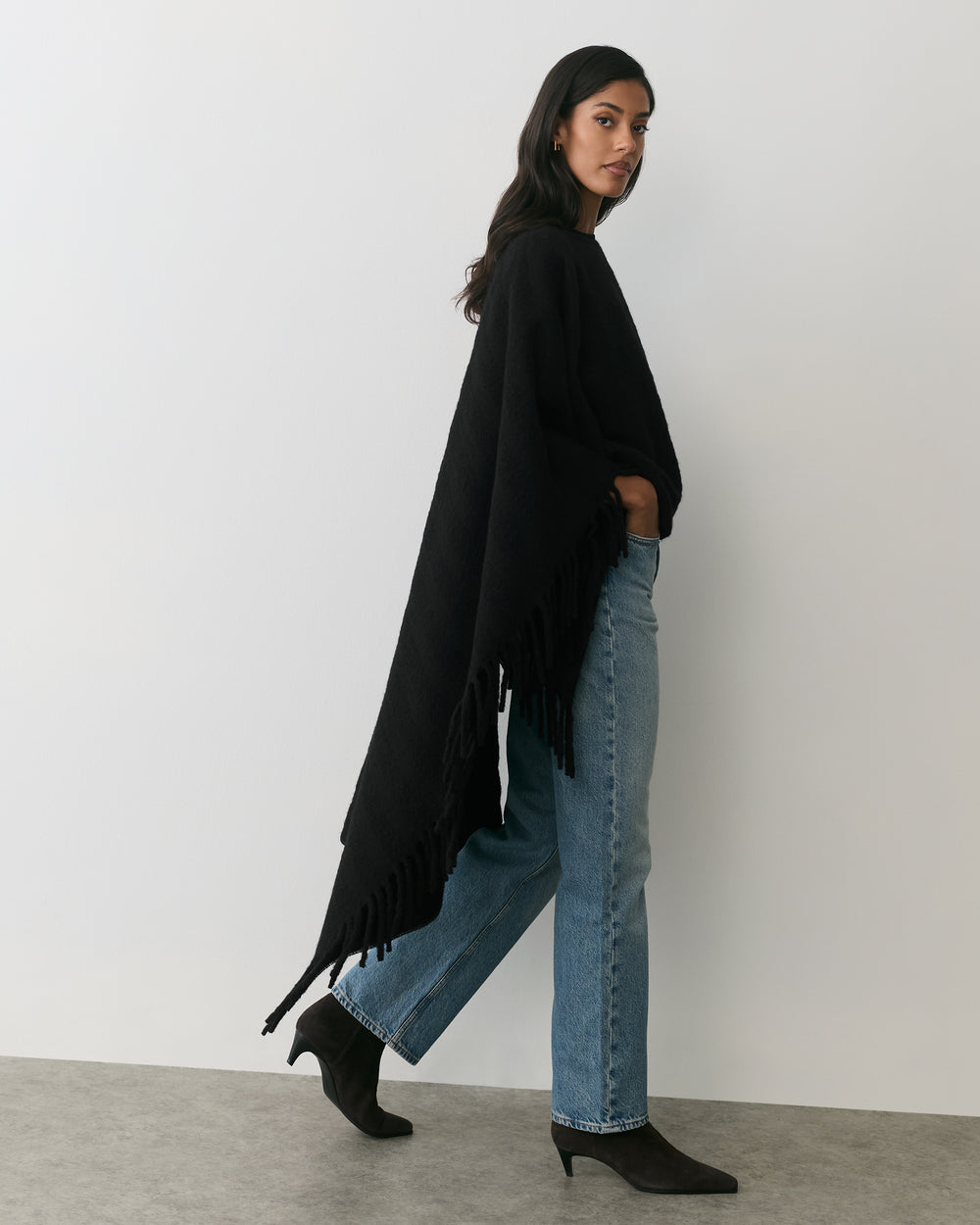 Person wearing cape, jeans, and ankle boots standing against wall.
