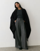 Person wearing a cape, long pants, and a long-sleeve shirt, standing against a wall.
