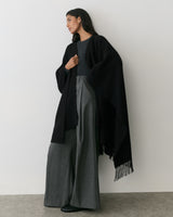 Person wearing a cape over a top and wide-leg trousers, standing against a plain wall.