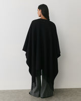 Person wearing a cape and wide-legged pants, standing against a plain wall.