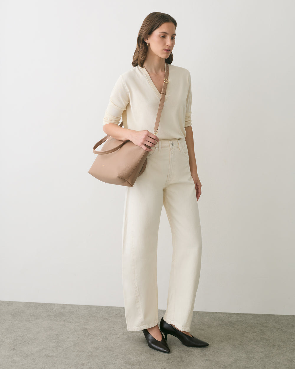 Person standing with crossbody bag wearing pants and long-sleeved top, looking to the side.