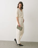Person standing with a small clutch, wearing a long-sleeve top and trousers.